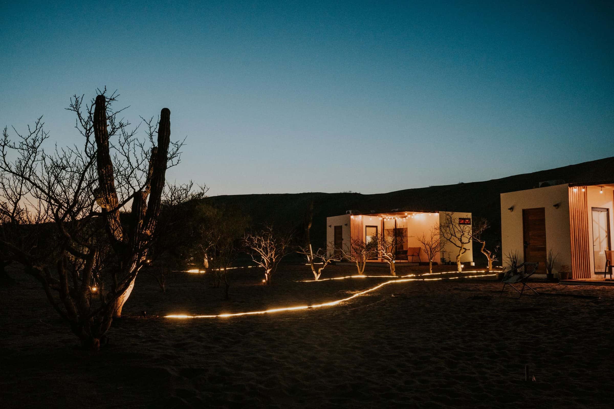 Two small desert homes at dusk, their porches lit, with a string of path lights across the sand.