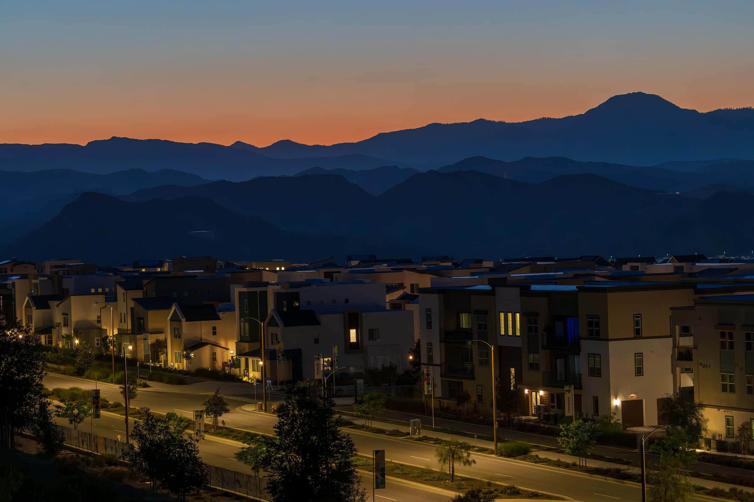 A residential neighbourhood at dusk, windows lit, with mountains behind it.